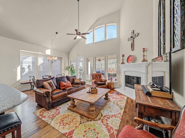 living room with ceiling fan with notable chandelier, a glass covered fireplace, high vaulted ceiling, and wood finished floors