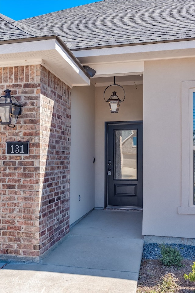 entrance to property featuring a shingled roof, brick siding, and stucco siding