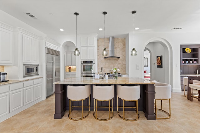 kitchen with arched walkways, visible vents, white cabinetry, wall chimney range hood, and built in appliances