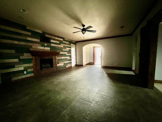 unfurnished living room featuring arched walkways, crown molding, a fireplace, ceiling fan, and baseboards