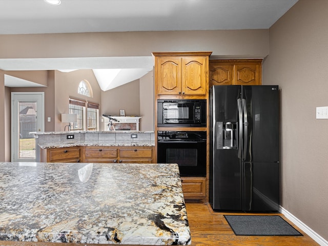 kitchen featuring light wood finished floors, baseboards, brown cabinetry, light stone counters, and black appliances