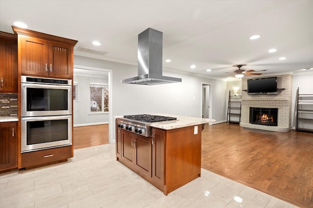 kitchen featuring visible vents, appliances with stainless steel finishes, a brick fireplace, island exhaust hood, and crown molding