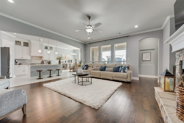 living area featuring ceiling fan with notable chandelier, a stone fireplace, dark wood-type flooring, and crown molding