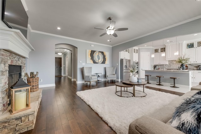 living room featuring arched walkways, ceiling fan, a stone fireplace, dark wood-style flooring, and baseboards