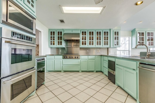 kitchen featuring stainless steel appliances, stainless steel countertops, a sink, green cabinetry, and under cabinet range hood