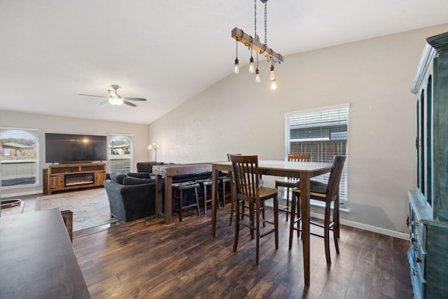 dining area featuring a warm lit fireplace, wood finished floors, a ceiling fan, baseboards, and vaulted ceiling