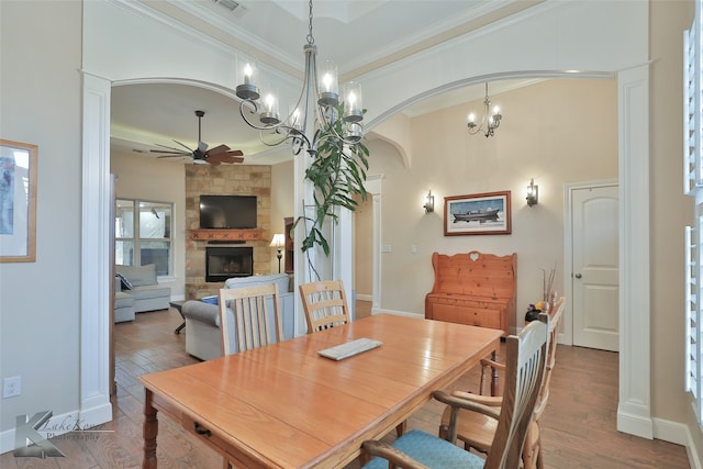 dining area featuring arched walkways, a stone fireplace, ceiling fan with notable chandelier, wood finished floors, and crown molding