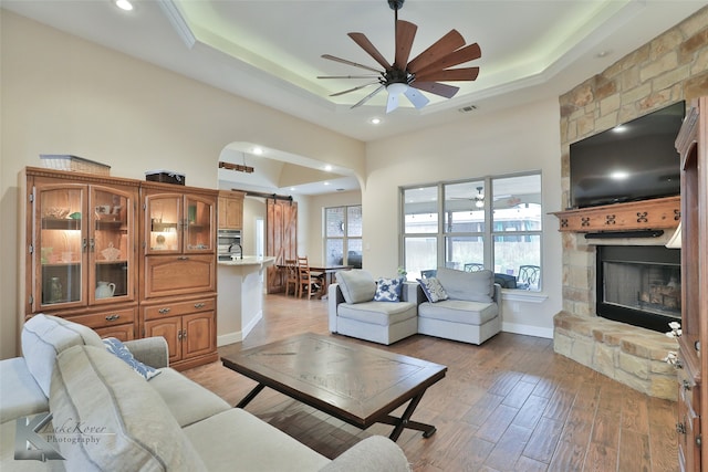 living area with light wood-type flooring, a raised ceiling, ceiling fan, and a stone fireplace