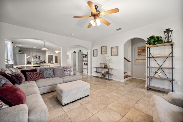 living room featuring light tile patterned floors, visible vents, arched walkways, ceiling fan, and stairway