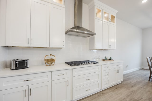 kitchen featuring light wood finished floors, decorative backsplash, wall chimney exhaust hood, stainless steel gas stovetop, and white cabinetry