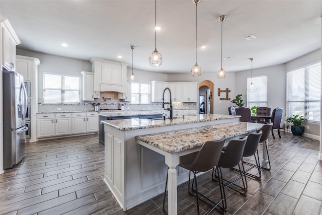 kitchen with arched walkways, a center island with sink, decorative backsplash, a sink, and stainless steel fridge with ice dispenser