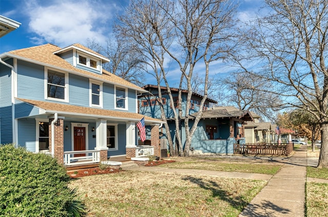 american foursquare style home featuring covered porch, roof with shingles, brick siding, and a front lawn