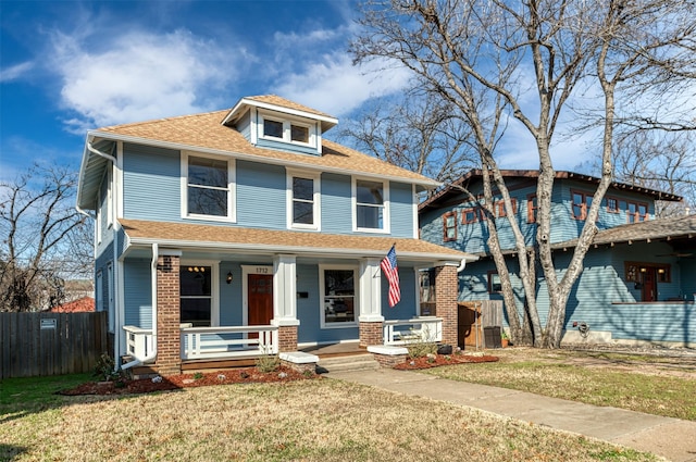 american foursquare style home with covered porch, a front lawn, a shingled roof, and fence