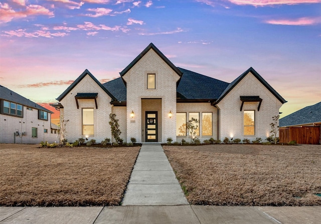 french country style house featuring a shingled roof, brick siding, and fence