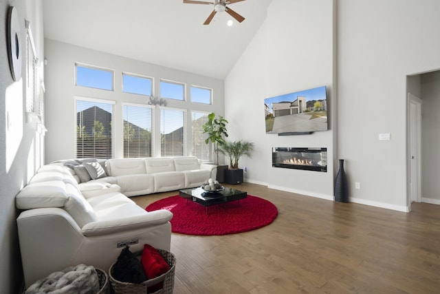 living area with baseboards, a ceiling fan, a glass covered fireplace, wood finished floors, and high vaulted ceiling