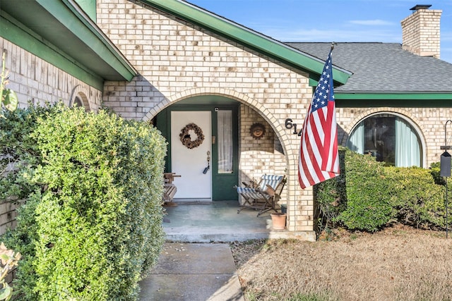 view of exterior entry featuring a shingled roof, brick siding, and a chimney