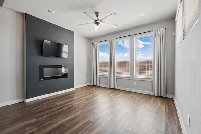 unfurnished living room featuring ceiling fan, baseboards, a glass covered fireplace, and dark wood-style floors