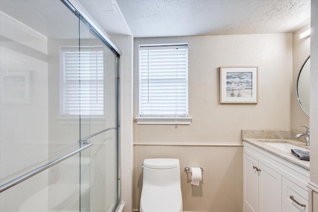 full bath with a textured ceiling, toilet, vanity, wainscoting, and a stall shower