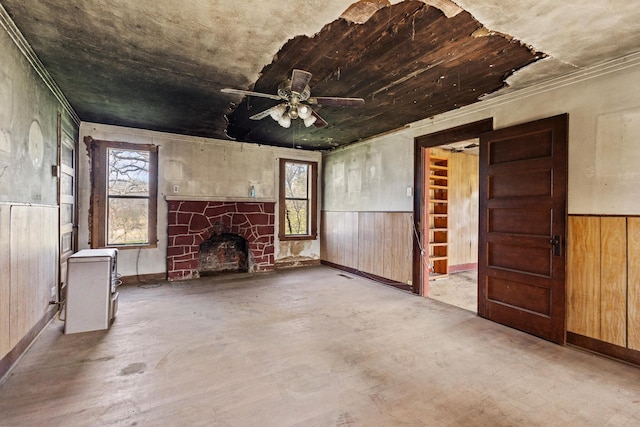 unfurnished living room featuring a wainscoted wall, ceiling fan, wooden walls, and a stone fireplace