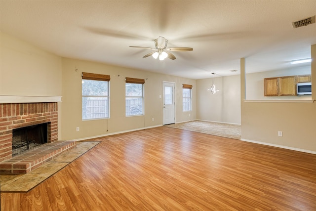 unfurnished living room with visible vents, light wood-style floors, a brick fireplace, baseboards, and ceiling fan with notable chandelier