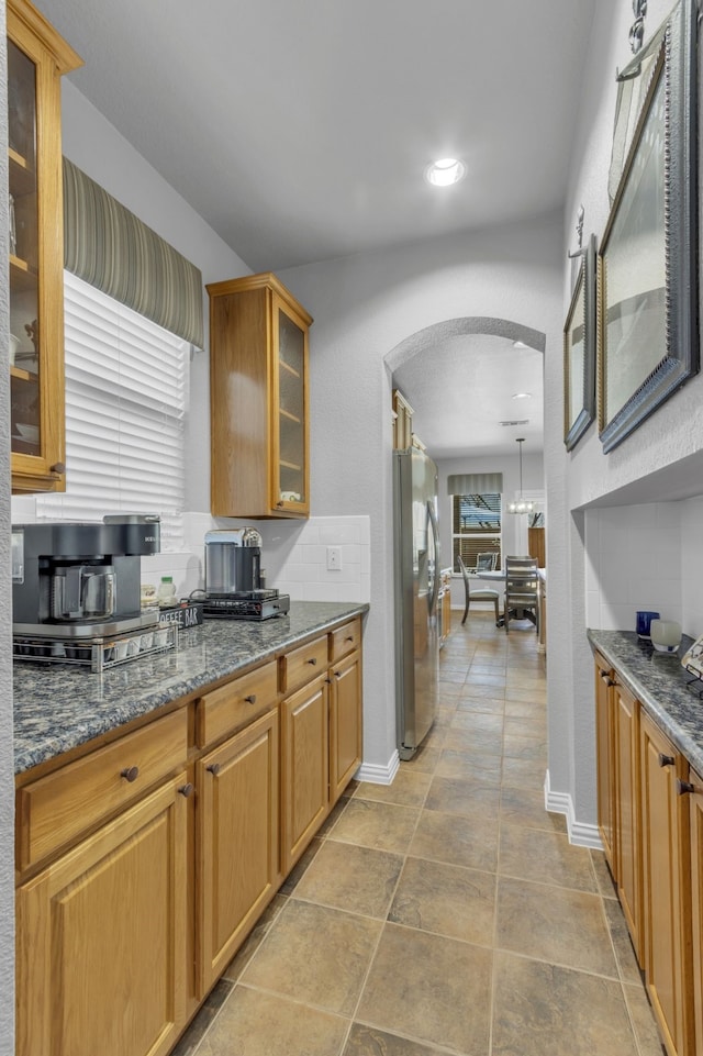 kitchen featuring arched walkways, baseboards, freestanding refrigerator, dark stone countertops, and glass insert cabinets