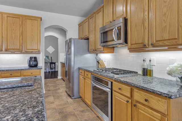 kitchen featuring appliances with stainless steel finishes, arched walkways, backsplash, and dark stone countertops