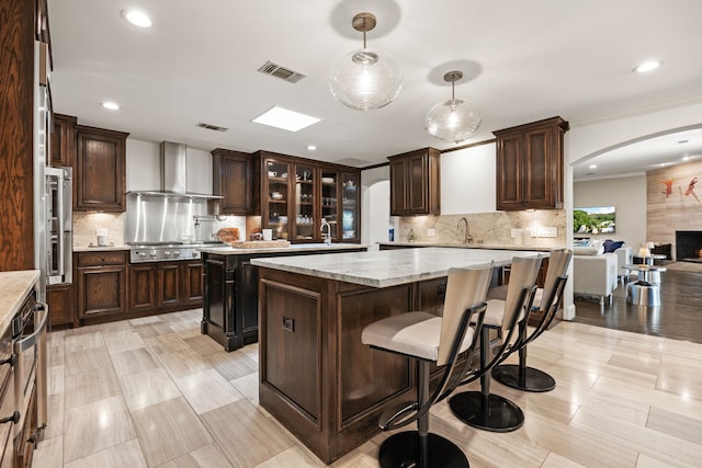 kitchen with arched walkways, dark brown cabinetry, visible vents, a center island, and wall chimney exhaust hood