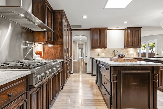 kitchen featuring arched walkways, wall chimney exhaust hood, dark brown cabinets, and stainless steel gas cooktop