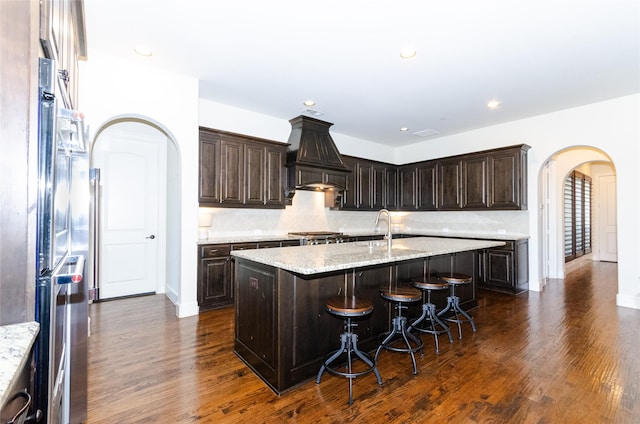 kitchen featuring dark wood-style floors, arched walkways, custom exhaust hood, a kitchen island with sink, and dark brown cabinetry