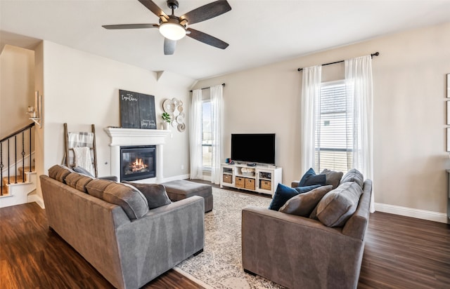 living room featuring ceiling fan, dark wood finished floors, baseboards, stairs, and a glass covered fireplace