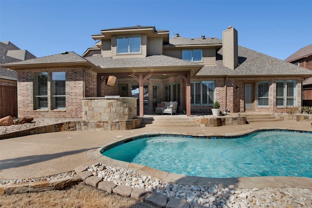 rear view of property featuring brick siding, roof with shingles, a chimney, an outdoor pool, and a patio