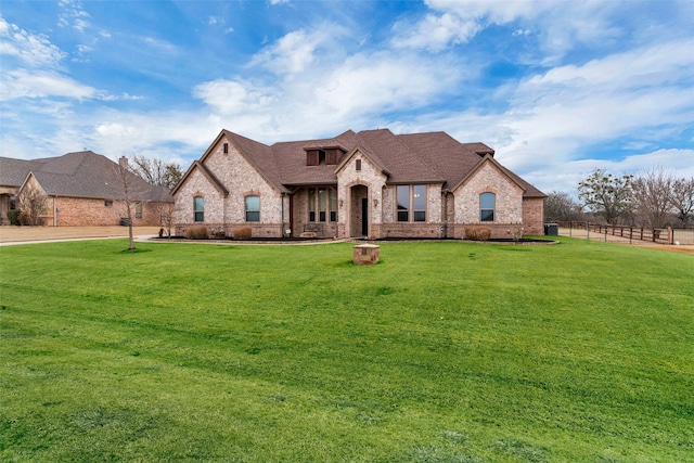 french country inspired facade with a shingled roof, a front yard, brick siding, and fence