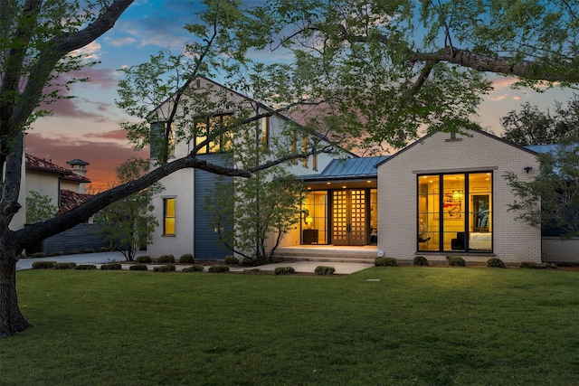 back of house at dusk with a standing seam roof, metal roof, a lawn, and brick siding