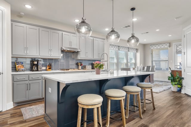 kitchen featuring dark wood-style floors, tasteful backsplash, gray cabinetry, an island with sink, and under cabinet range hood