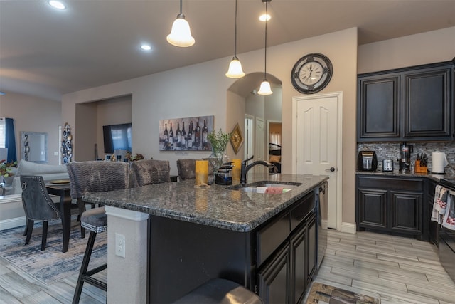 kitchen featuring a center island with sink, decorative backsplash, a sink, dark stone countertops, and a kitchen bar