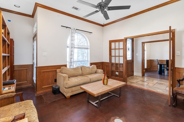 living area featuring a wainscoted wall, crown molding, visible vents, a ceiling fan, and concrete floors