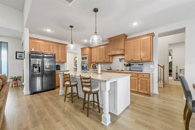 kitchen with light wood-style floors, a kitchen island with sink, stainless steel oven, fridge with ice dispenser, and premium range hood