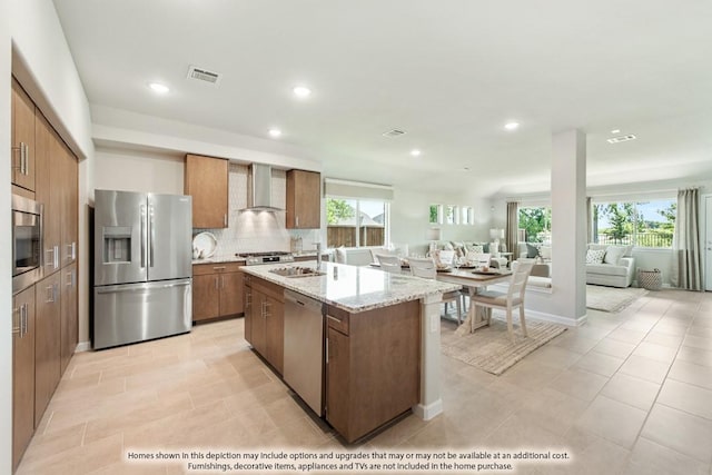 kitchen with stainless steel appliances, backsplash, brown cabinetry, open floor plan, and wall chimney exhaust hood