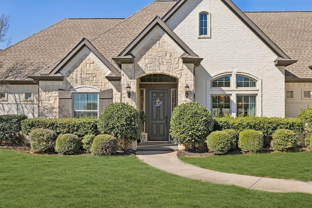 french country style house featuring stone siding, a shingled roof, a front lawn, and brick siding