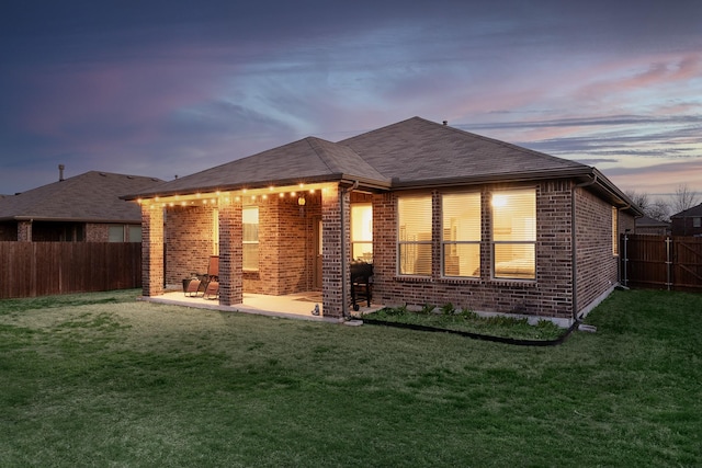 back of house at dusk featuring a yard, a fenced backyard, a patio, and brick siding