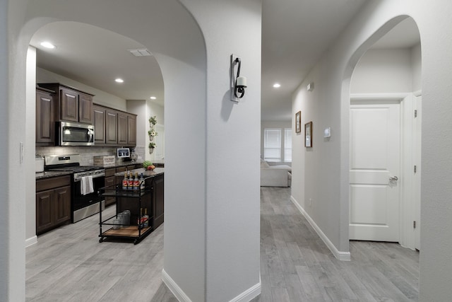 kitchen with arched walkways, dark brown cabinetry, stainless steel appliances, light wood-style floors, and backsplash