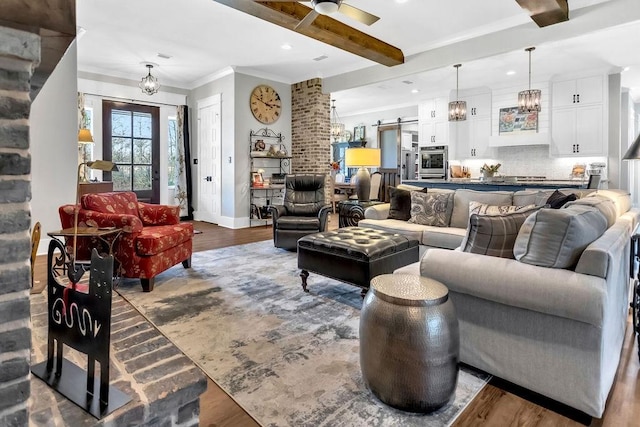 living area featuring a barn door, dark wood finished floors, ornamental molding, beam ceiling, and ceiling fan with notable chandelier