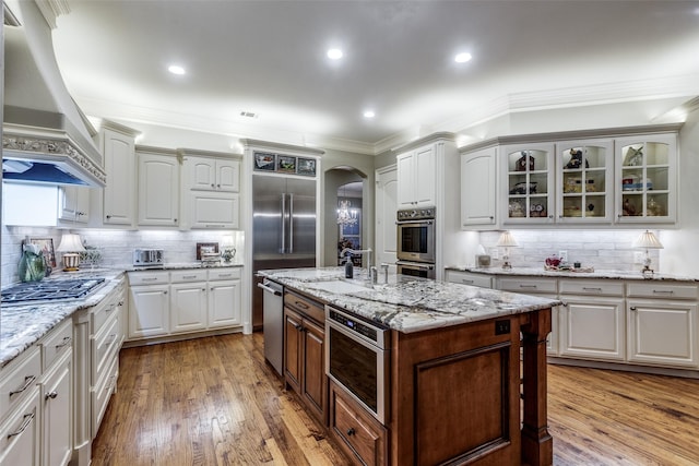 kitchen featuring arched walkways, custom range hood, wood-type flooring, glass insert cabinets, and crown molding