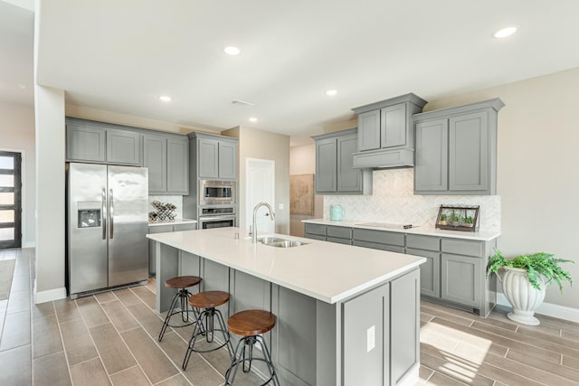 kitchen featuring stainless steel appliances, a sink, a center island with sink, and gray cabinetry