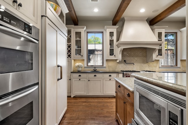 kitchen featuring dark wood-type flooring, appliances with stainless steel finishes, tasteful backsplash, beamed ceiling, and custom range hood