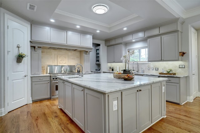 kitchen featuring light stone counters, a sink, light wood finished floors, an island with sink, and a raised ceiling