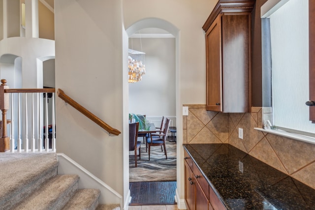 kitchen with arched walkways, a notable chandelier, backsplash, dark stone counters, and brown cabinetry