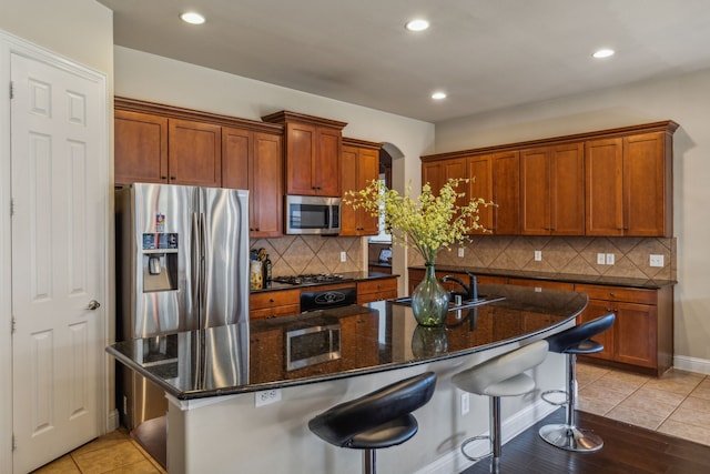 kitchen featuring light tile patterned floors, dark stone counters, an island with sink, stainless steel appliances, and a sink