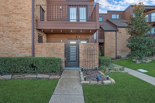 view of exterior entry featuring a balcony, brick siding, fence, a lawn, and a gate