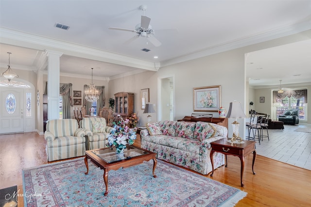 living area featuring decorative columns, visible vents, ornamental molding, light wood-style floors, and ceiling fan with notable chandelier